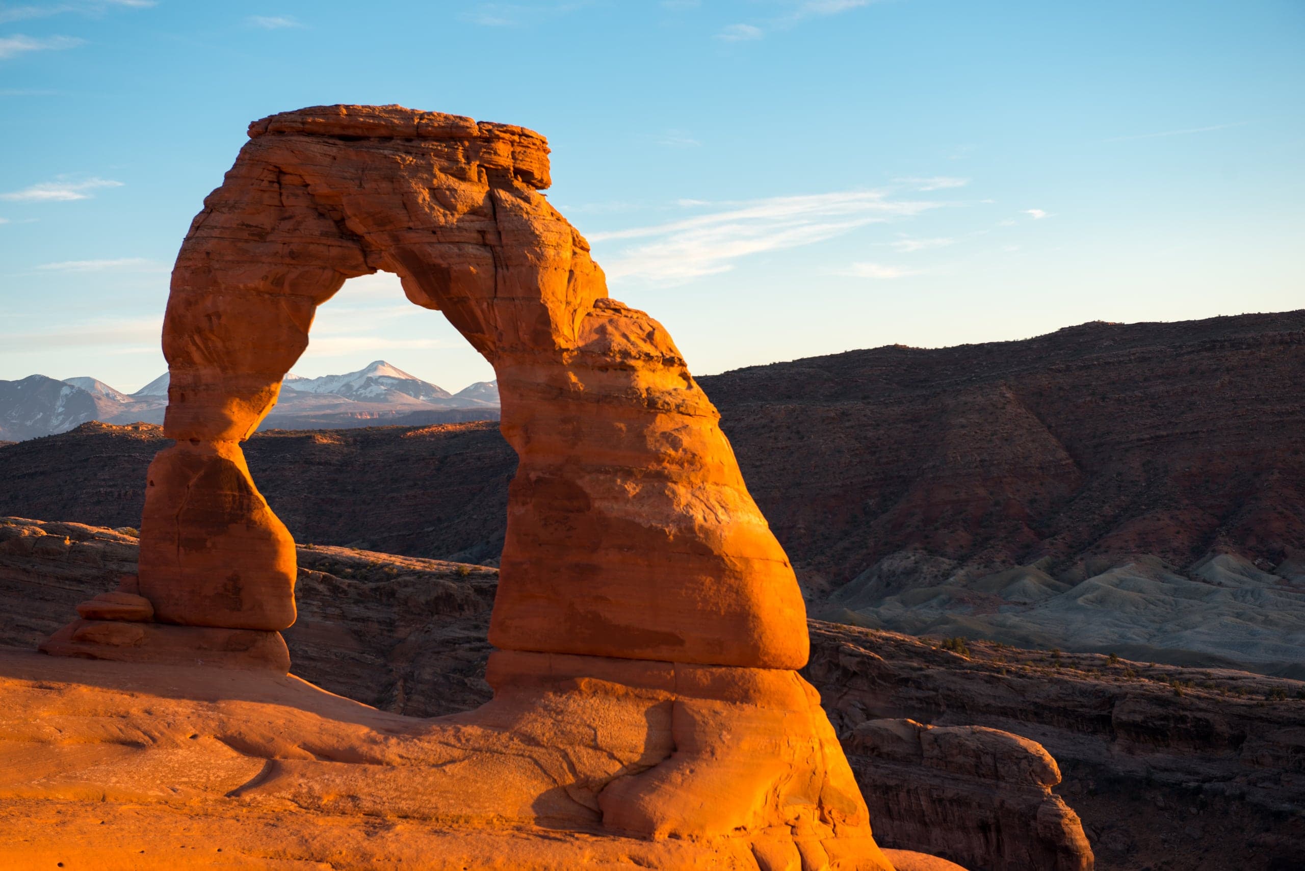 Delicate Arch, a free-standing sandstone arch glowing orange in low sunlight, stands on bare slickrock. Snow-dusted mountains are visible through the opening beneath a pale blue sky.