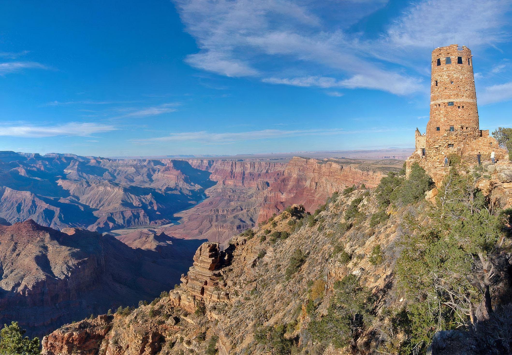 The circular stone Desert View Watchtower stands on the canyon rim at the right of the frame. To the left, the layered red and tan cliffs and terraces of the Grand Canyon recede into haze under a deep blue sky.